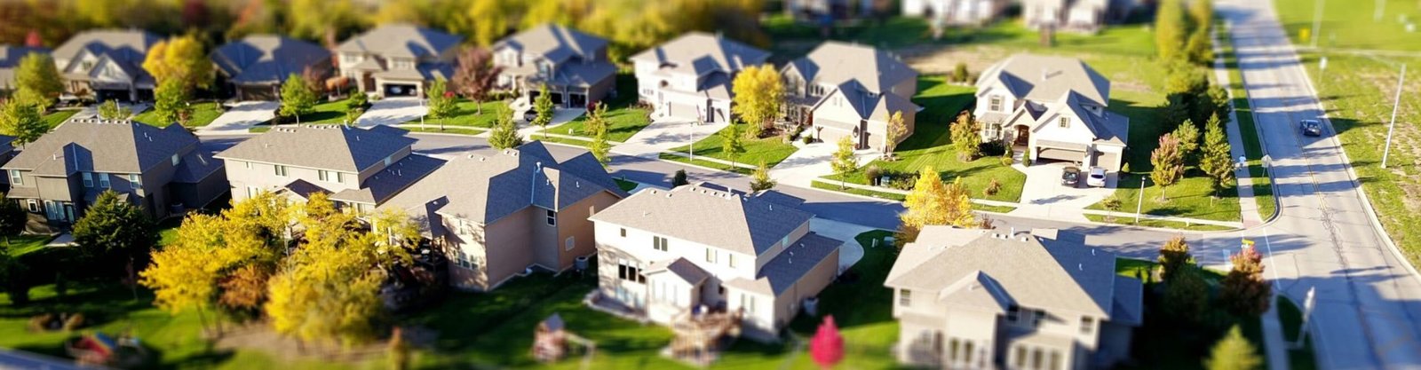 Tilt-shift aerial photo of a suburban neighborhood with green lawns and roads.
